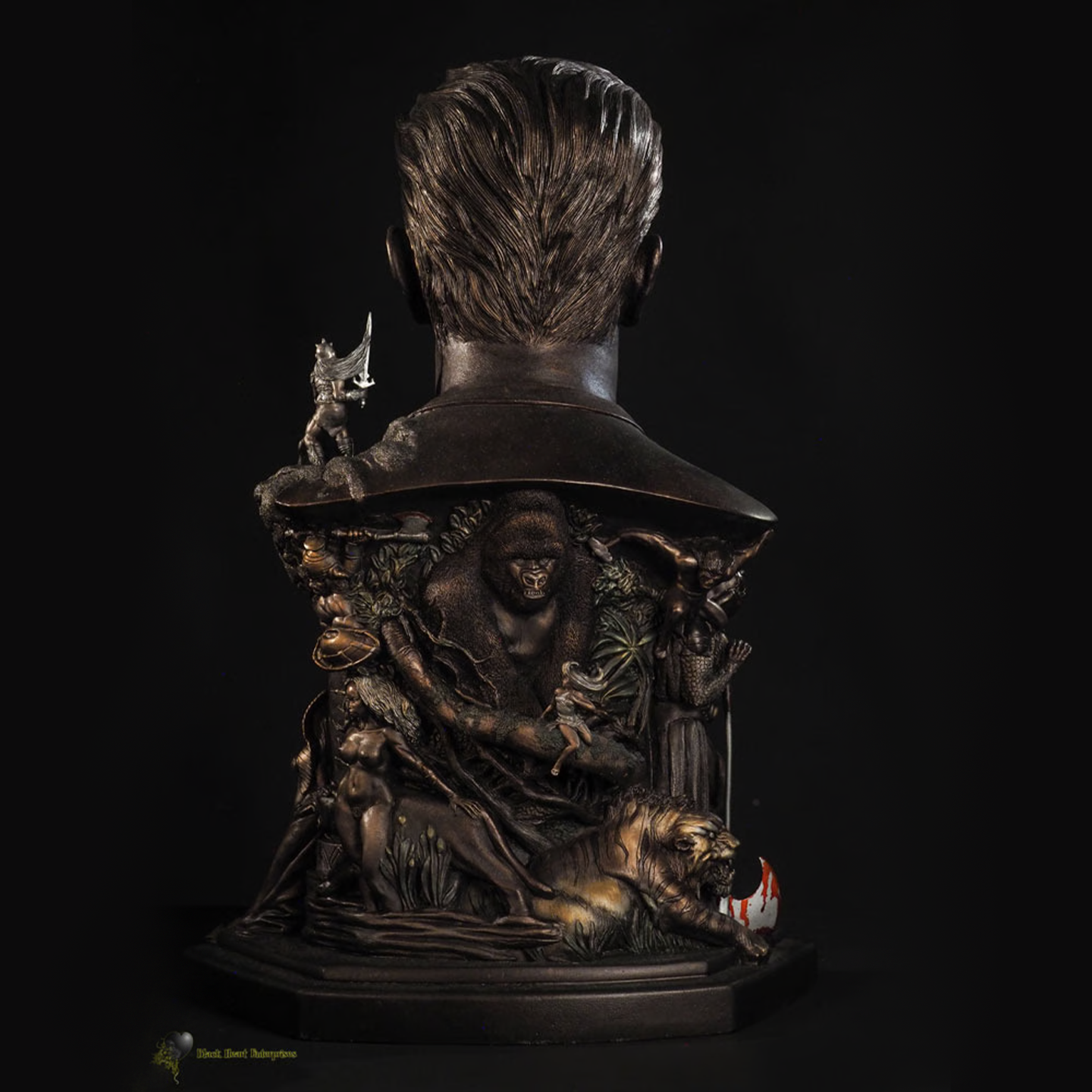 The back of a faux bronze polystone bust of Frank Frazetta, featuring intricate bas-relief sculptures of his iconic fantasy and adventure artwork. The design includes a detailed jungle scene with a massive gorilla, a fierce tiger, and dynamic figures of warriors and heroines in battle. The highly detailed craftsmanship captures Frazetta’s legendary artistry, bringing his creations to life in a striking tribute.