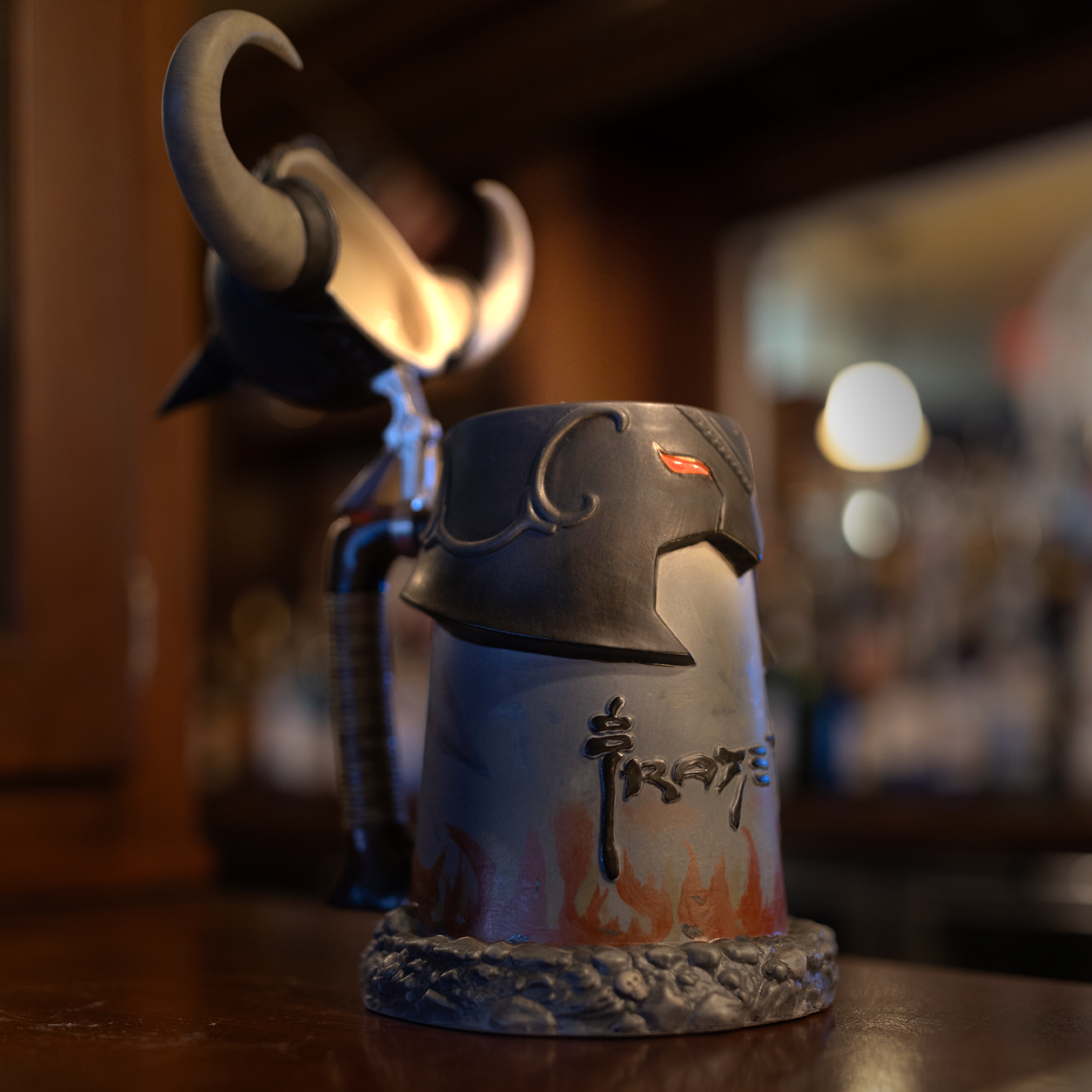 A vintage Death Dealer stein sits open on a polished wooden bar, illuminated by warm ambient lighting. The ceramic stein features a sculpted warrior helmet with curved horns, glowing red eyes, and intricate engravings. The base is adorned with a textured stone-like design and fiery artwork, with "Frazetta" inscribed on the front. A blurred background of liquor bottles and bar lights adds depth to the scene.