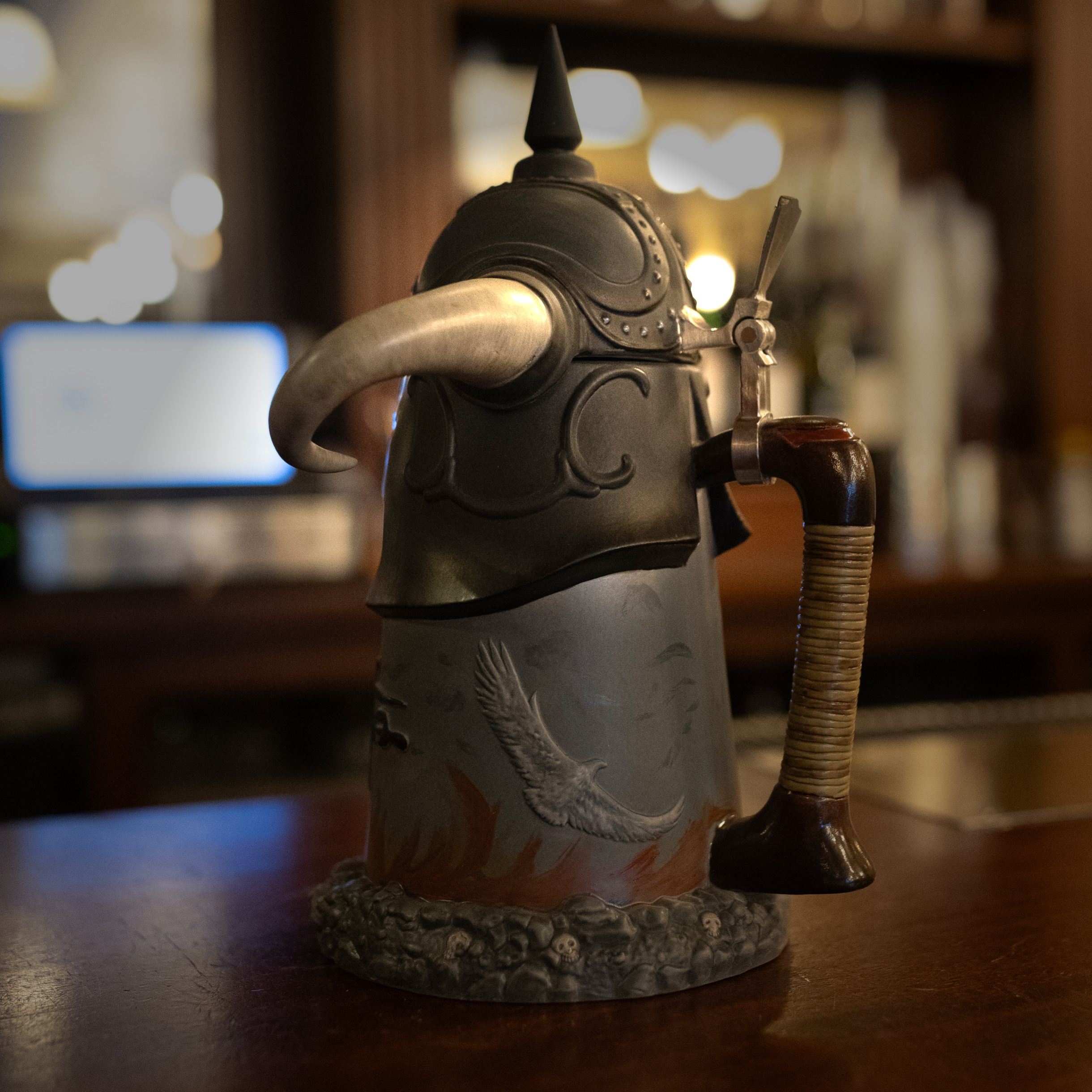 A vintage Death Dealer stein sits on a polished wooden bar, illuminated by warm ambient lighting. The ceramic stein features a sculpted warrior helmet with curved horns, glowing red eyes, and intricate engravings. The base is adorned with a textured stone-like design and fiery artwork. A blurred background of liquor bottles and bar lights adds depth to the scene.