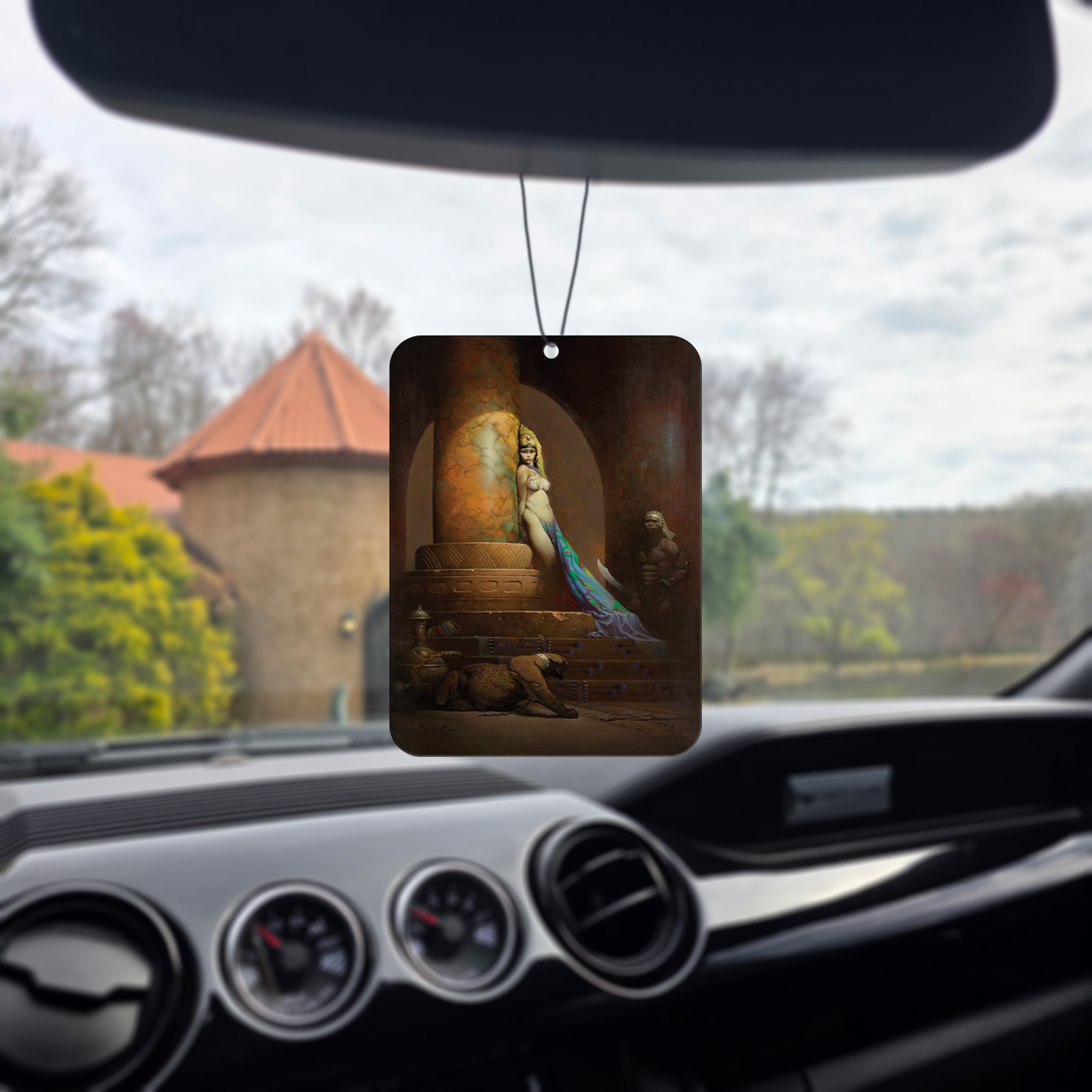 Frazetta Egyptian Queen air freshener hanging in a car with black interior. The Frazetta Art Museum sits in the background with gloomy clouds overhead.