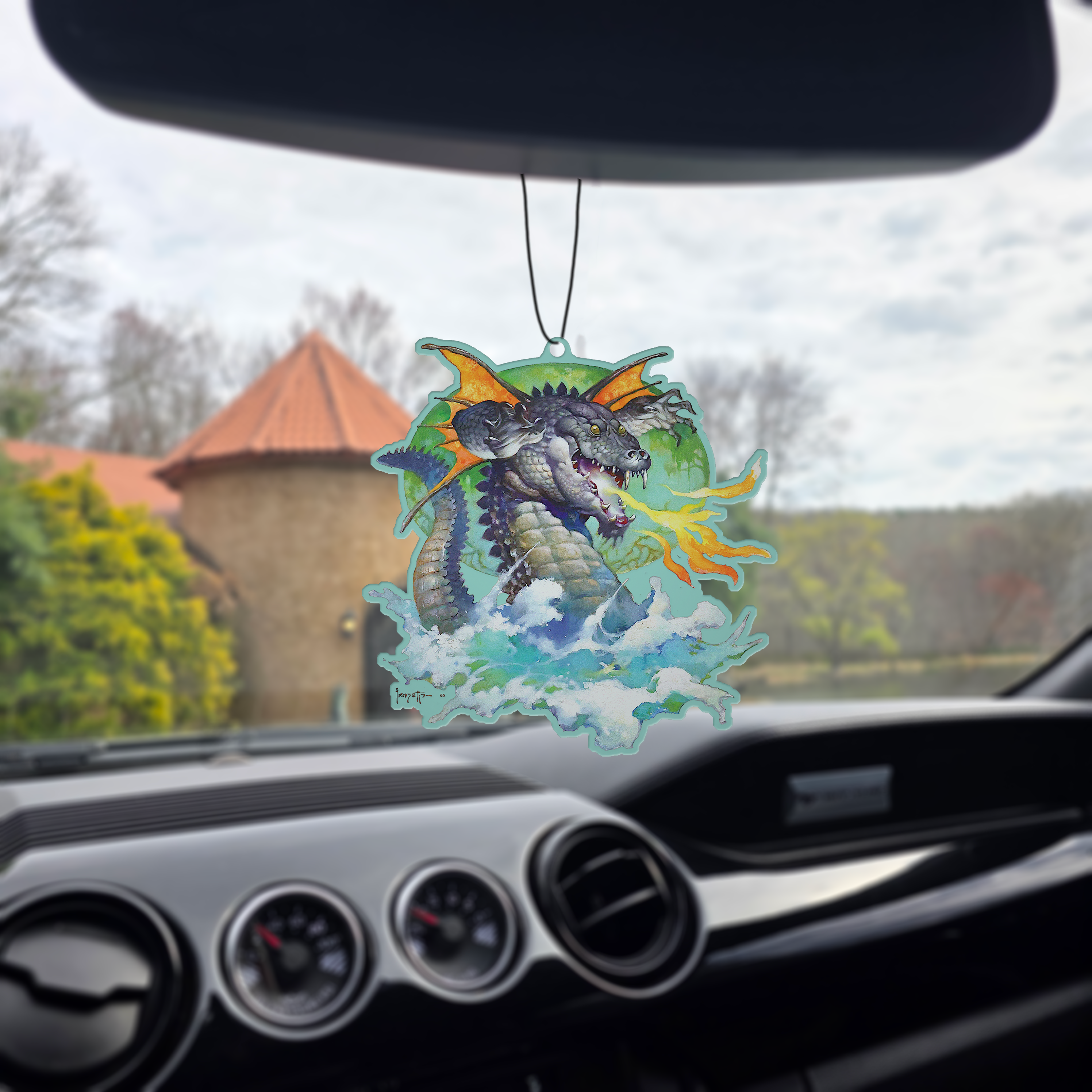 Frazetta Flying Alligator air freshener hanging in a car with black interior. The Frazetta Art Museum sits in the background with gloomy clouds overhead.