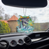 Frazetta Flying Alligator air freshener hanging in a car with black interior. The Frazetta Art Museum sits in the background with gloomy clouds overhead.