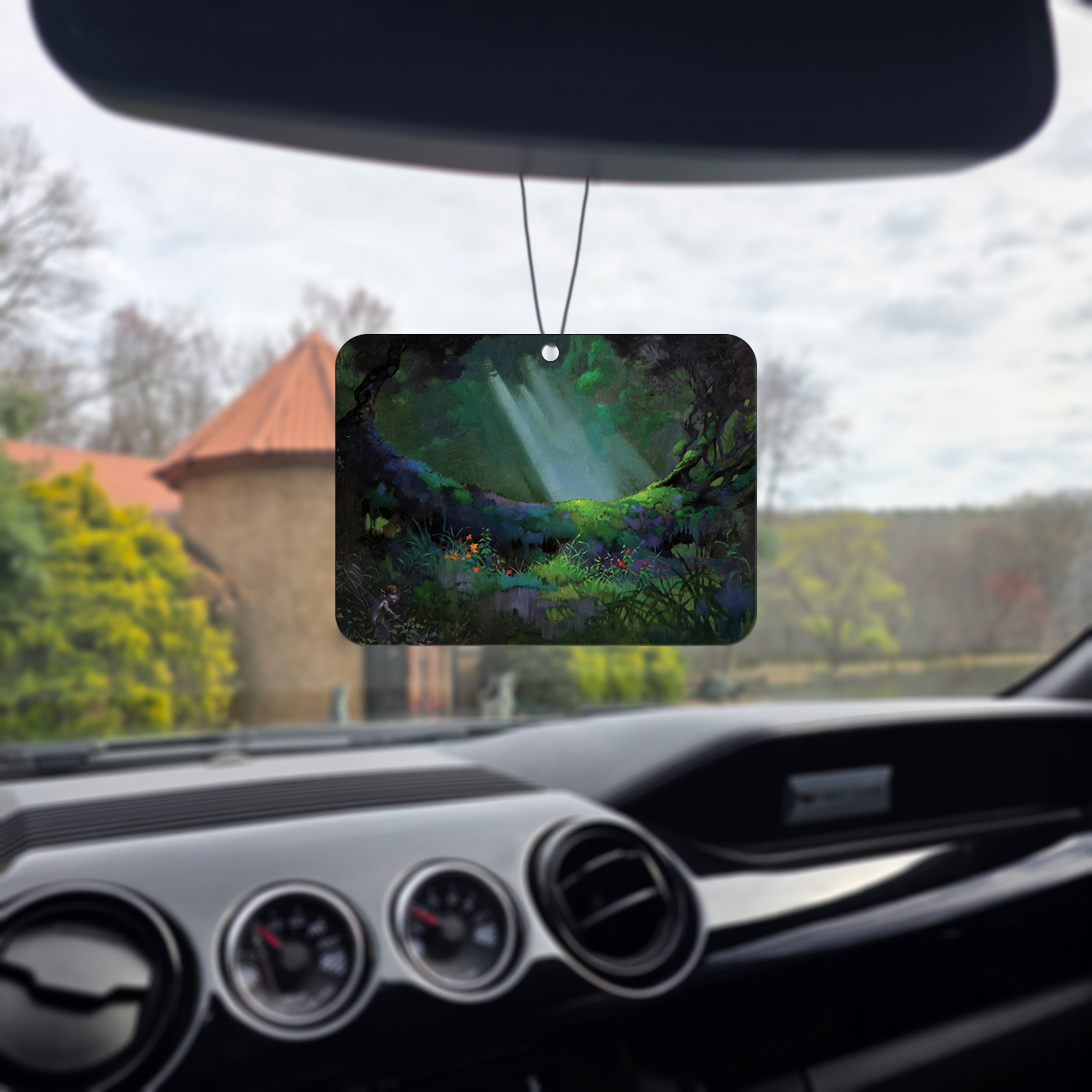 Frazetta Glade of the Pixies air freshener hanging in a car with black interior. The Frazetta Art Museum sits in the background with gloomy clouds overhead.