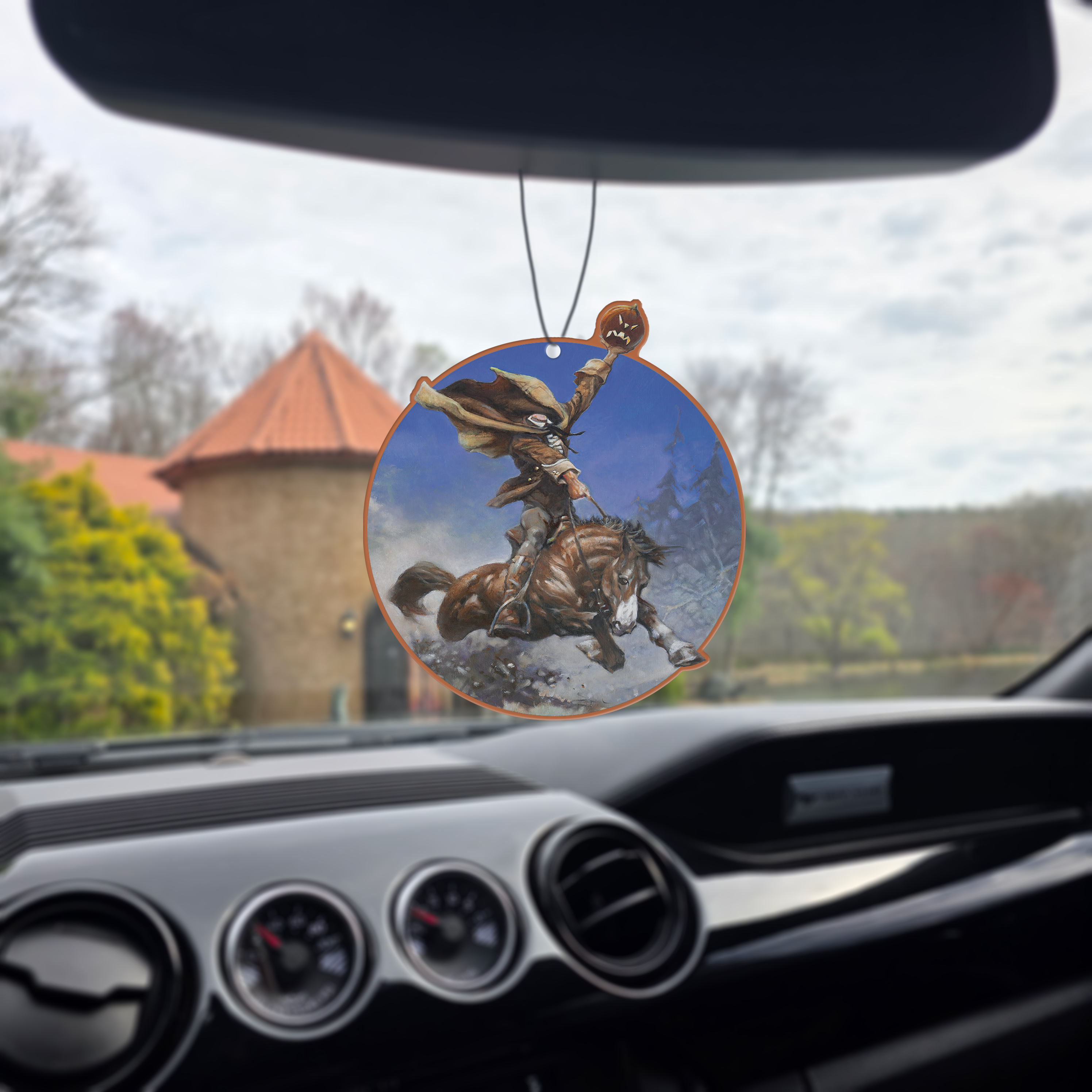 Frazetta Headless Horseman air freshener hanging in a car with black interior. The Frazetta Art Museum sits in the background with gloomy clouds overhead.