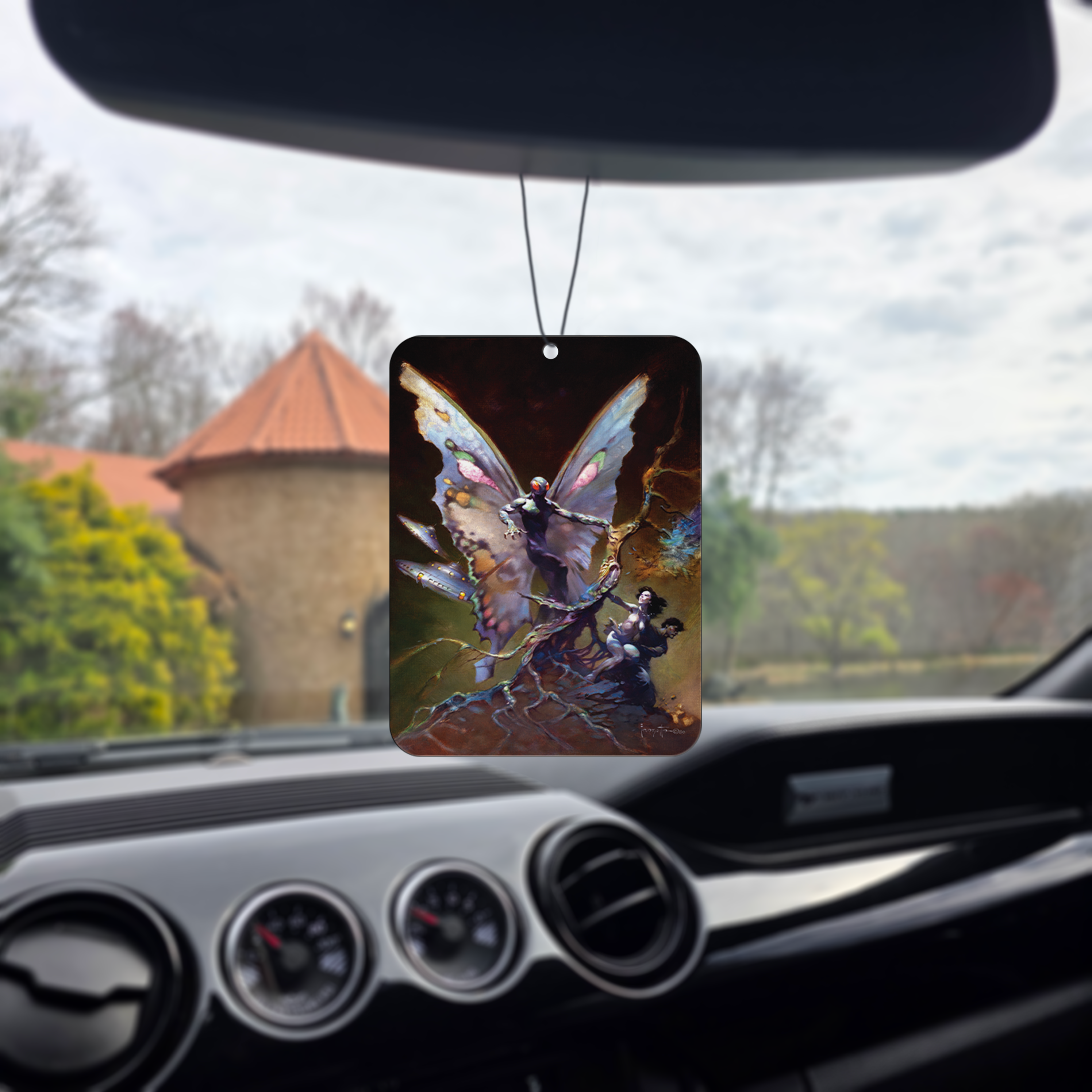 Photo of the Frazetta Mothman air freshener hanging in a car with black interior. The Frazetta Art Museum sits in the background with gloomy clouds overhead.