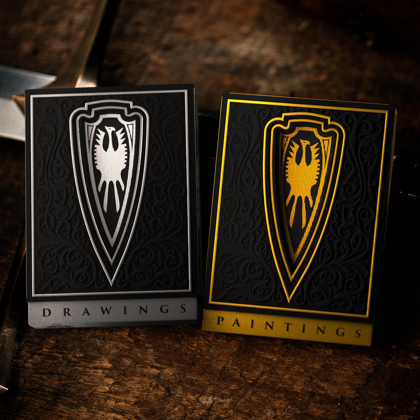 Two elegantly designed playing card decks rest on a rustic wooden surface. The "Drawings" deck, in black and silver, and the "Paintings" deck, in black and gold, feature intricate embossed detailing with a bold shield emblem at the center. The rich contrast of metallic accents against the dark background highlights the premium craftsmanship and collector’s appeal of the set.