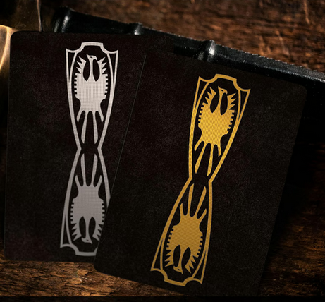 Two elegantly designed playing cards rest on a dark wooden surface, featuring a symmetrical shield emblem with a stylized eagle. One card has a gold-foil design, while the other features silver, both set against a rich black background.