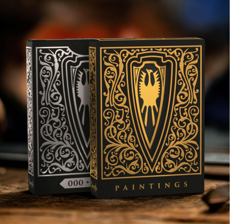 Two elegantly designed playing card decks rest on a wooden surface, featuring intricate gold and silver detailing. The black and gold deck prominently displays a shield emblem with a stylized eagle at its center, surrounded by ornate filigree, with "PAINTINGS" inscribed at the bottom. The black and silver deck, positioned slightly behind, mirrors the same intricate design, with a numbered label on the side. The background is softly blurred, creating a rich, atmospheric setting that highlights the luxurious