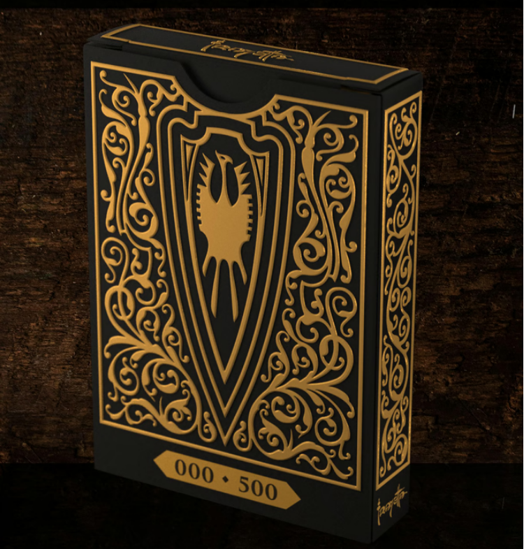 A luxurious black and gold playing card box featuring an intricate, ornate design. The front showcases a bold shield emblem with a stylized eagle at its center, surrounded by elegant filigree patterns. A numbered label at the bottom indicates a limited edition release (000/500). The box is set against a dark, textured wooden background, emphasizing its premium craftsmanship and collectible nature.