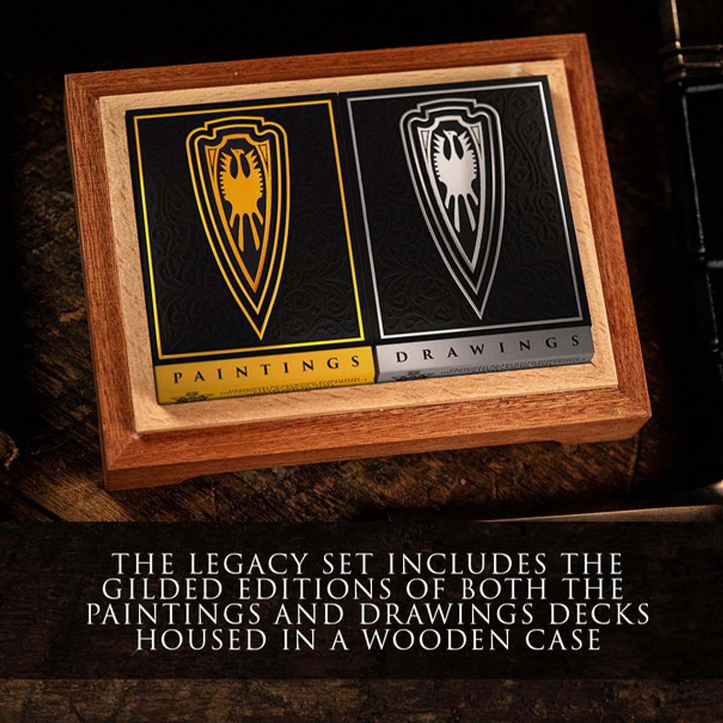 A luxurious wooden collector’s box holds two elegantly designed playing card decks from the Legacy Edition set. The "Paintings" deck, in black and gold, and the "Drawings" deck, in black and silver, feature intricate shield emblems and ornate detailing. The Frazetta logo is engraved on the inside of the wooden lid, adding to the premium presentation. The set rests on a rich wooden surface, surrounded by dark-toned accessories, emphasizing its collector’s appeal and craftsmanship.