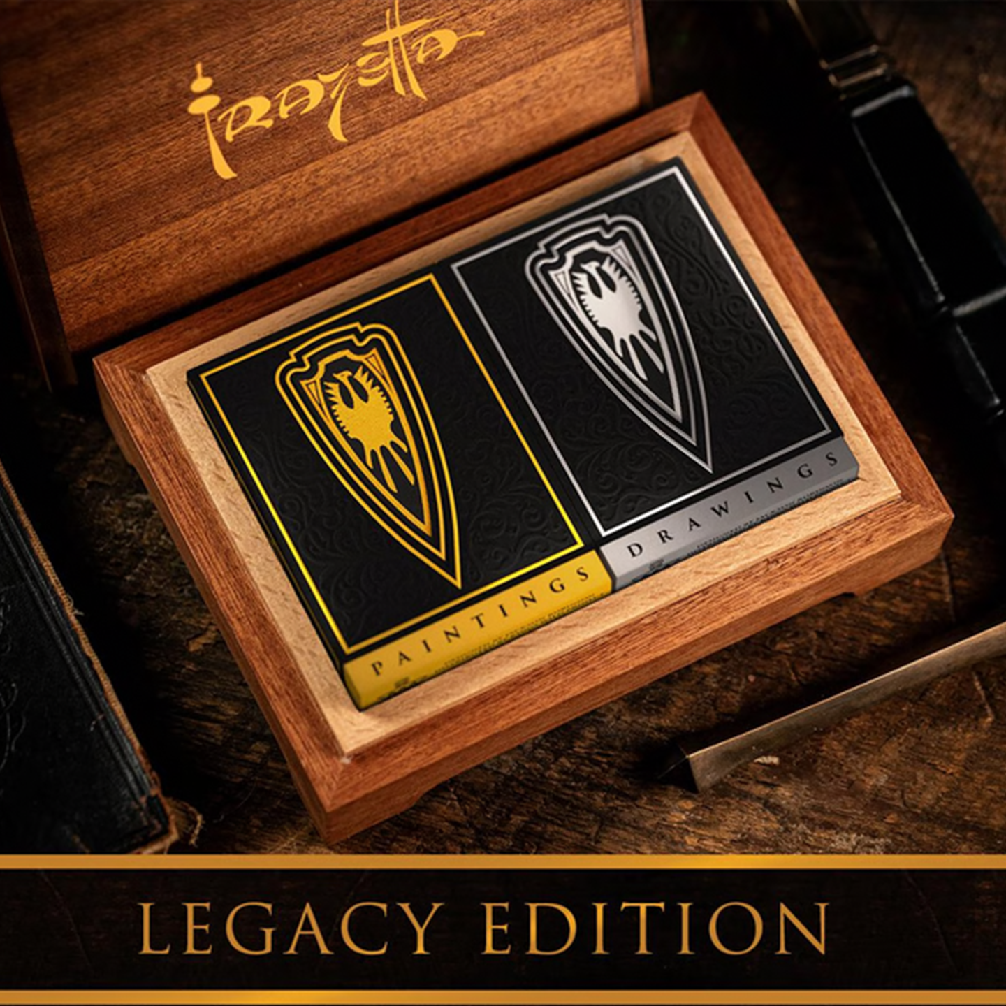 A luxurious wooden collector’s box holds two elegantly designed playing card decks from the Legacy Edition set. The "Paintings" deck, in black and gold, and the "Drawings" deck, in black and silver, feature intricate shield emblems and ornate detailing. The Frazetta logo is engraved on the inside of the wooden lid, adding to the premium presentation. The set rests on a rich wooden surface, surrounded by dark-toned accessories, emphasizing its collector’s appeal and craftsmanship.