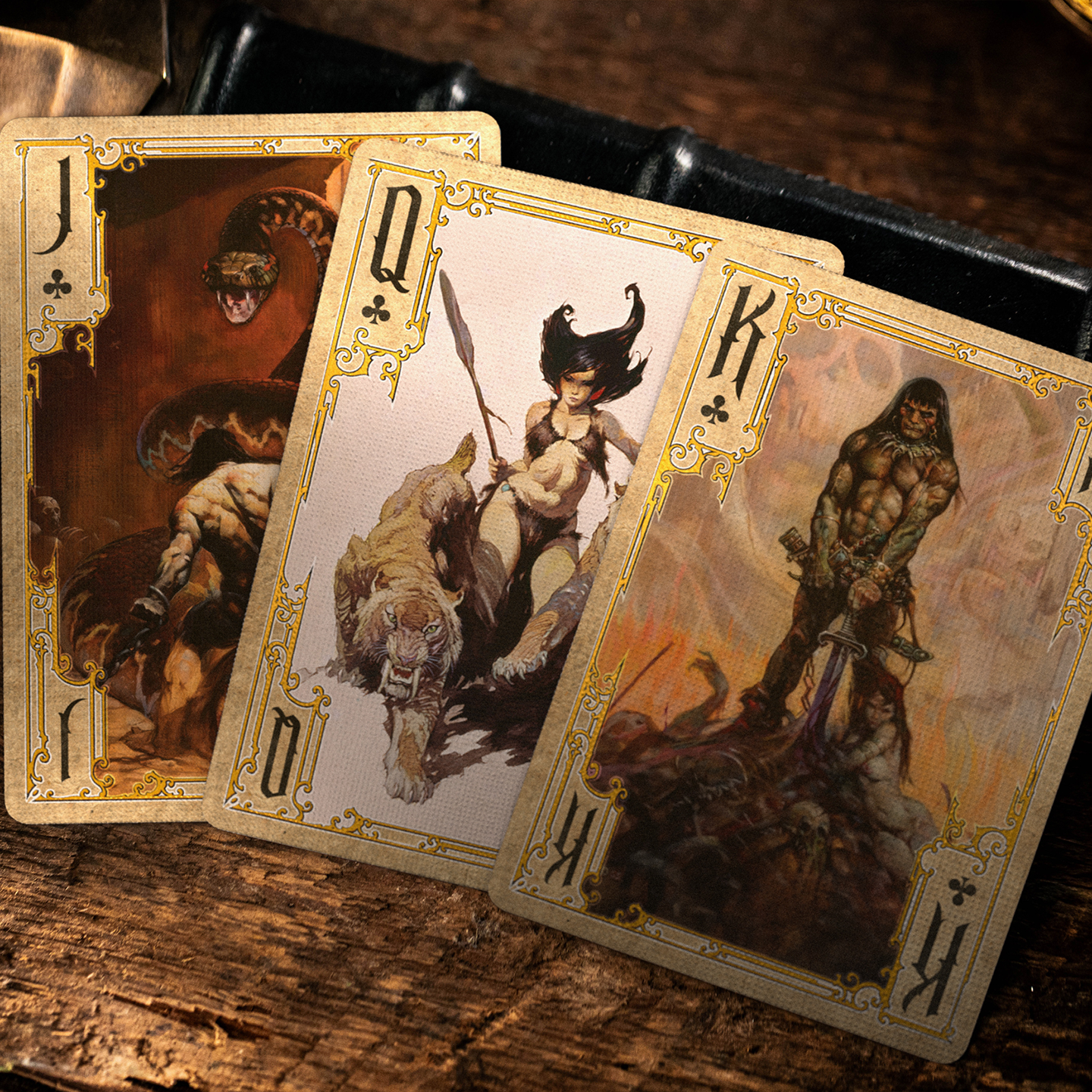 A close-up of a King, Queen, and Jack of clubs from the Frazetta Paintings Edition playing card deck. The cards feature an ornate, vintage-style border with delicate filigree and a stylized club symbol at its center. Beneath it, additional cards are partially visible, showcasing intricate artwork and embossed detailing. A knife rests nearby, enhancing the dramatic composition.