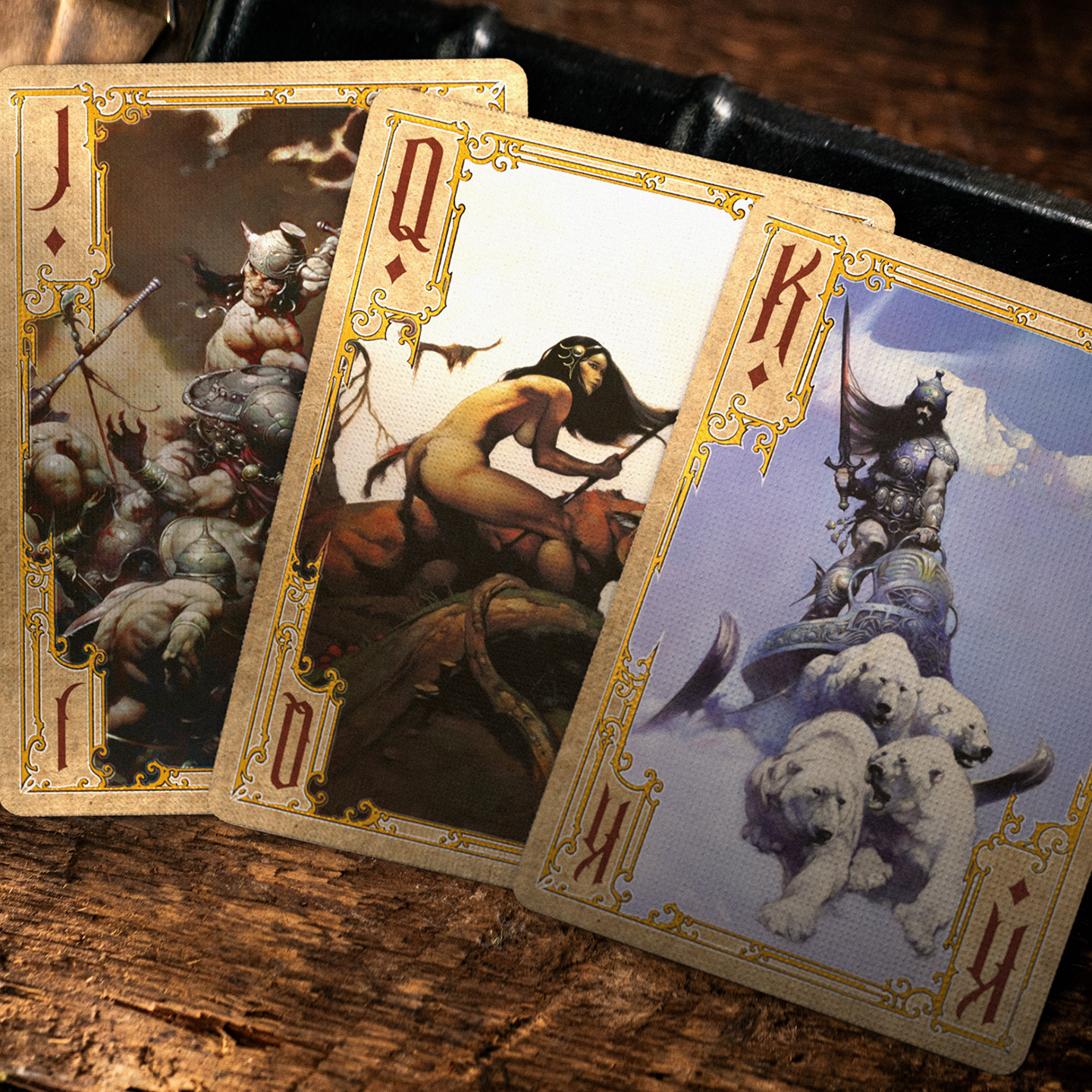 A close-up of a King, Queen, and Jack of Diamonds from the Frazetta Paintings Edition playing card deck. The cards feature an ornate, vintage-style border with delicate filigree and a stylized club symbol at its center. Beneath it, additional cards are partially visible, showcasing intricate artwork and embossed detailing. A knife rests nearby, enhancing the dramatic composition.