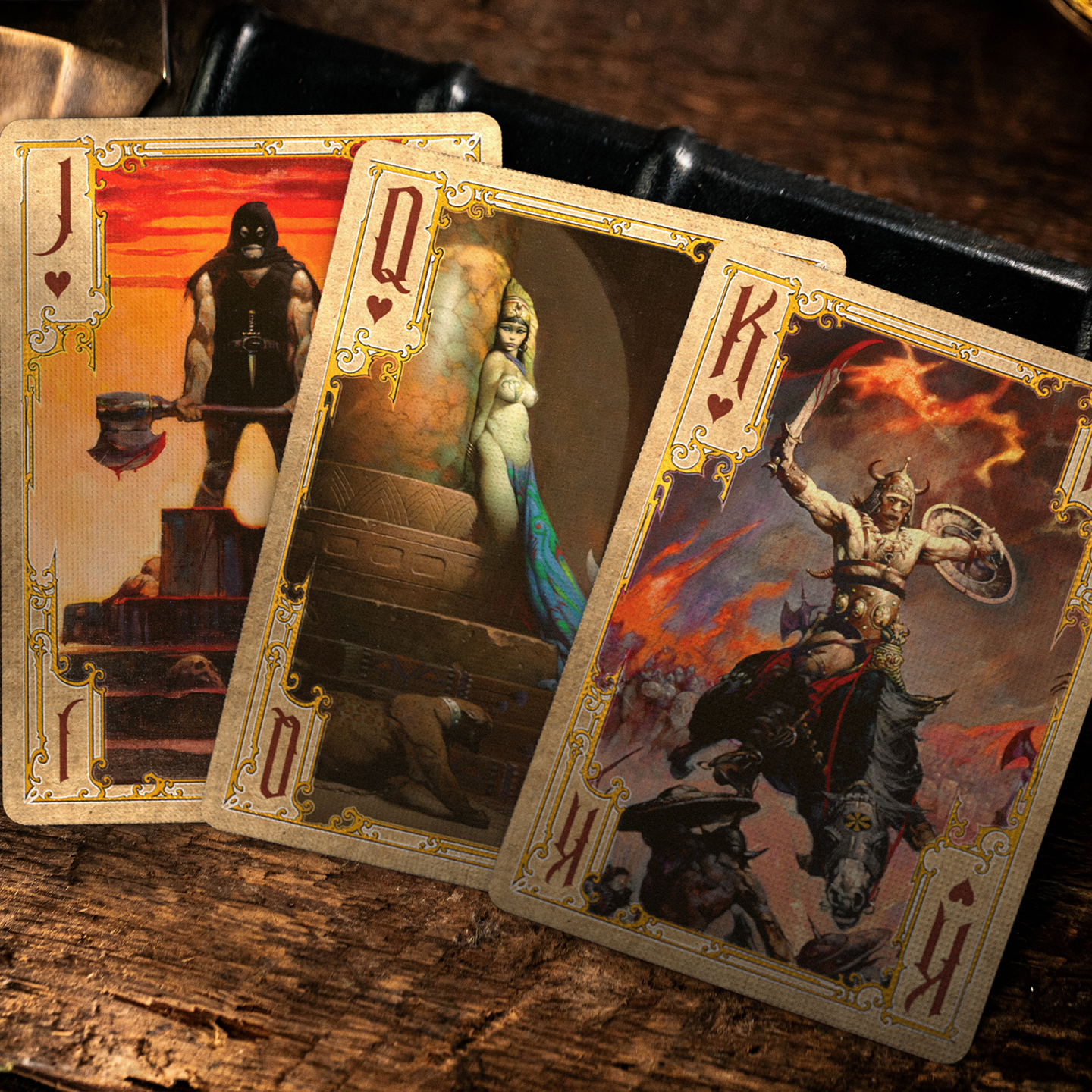 A close-up of a King, Queen, and Jack of Hearts from the Frazetta Paintings Edition playing card deck. The cards feature an ornate, vintage-style border with delicate filigree and a stylized club symbol at its center. Beneath it, additional cards are partially visible, showcasing intricate artwork and embossed detailing. A knife rests nearby, enhancing the dramatic composition.