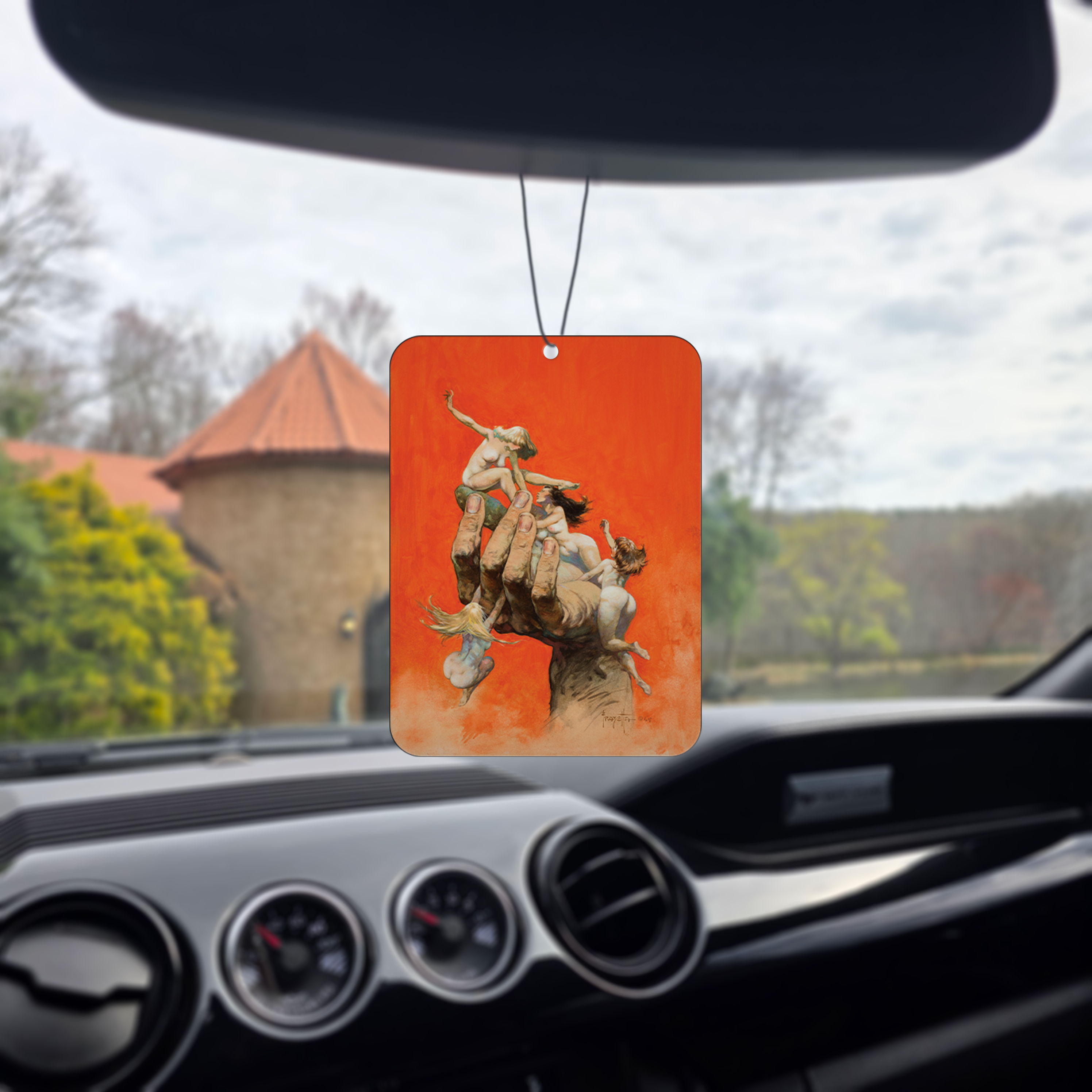 Photo of the Frazetta Reassembled Man air freshener hanging in a car with black interior. The Frazetta Art Museum sits in the background with gloomy clouds overhead.