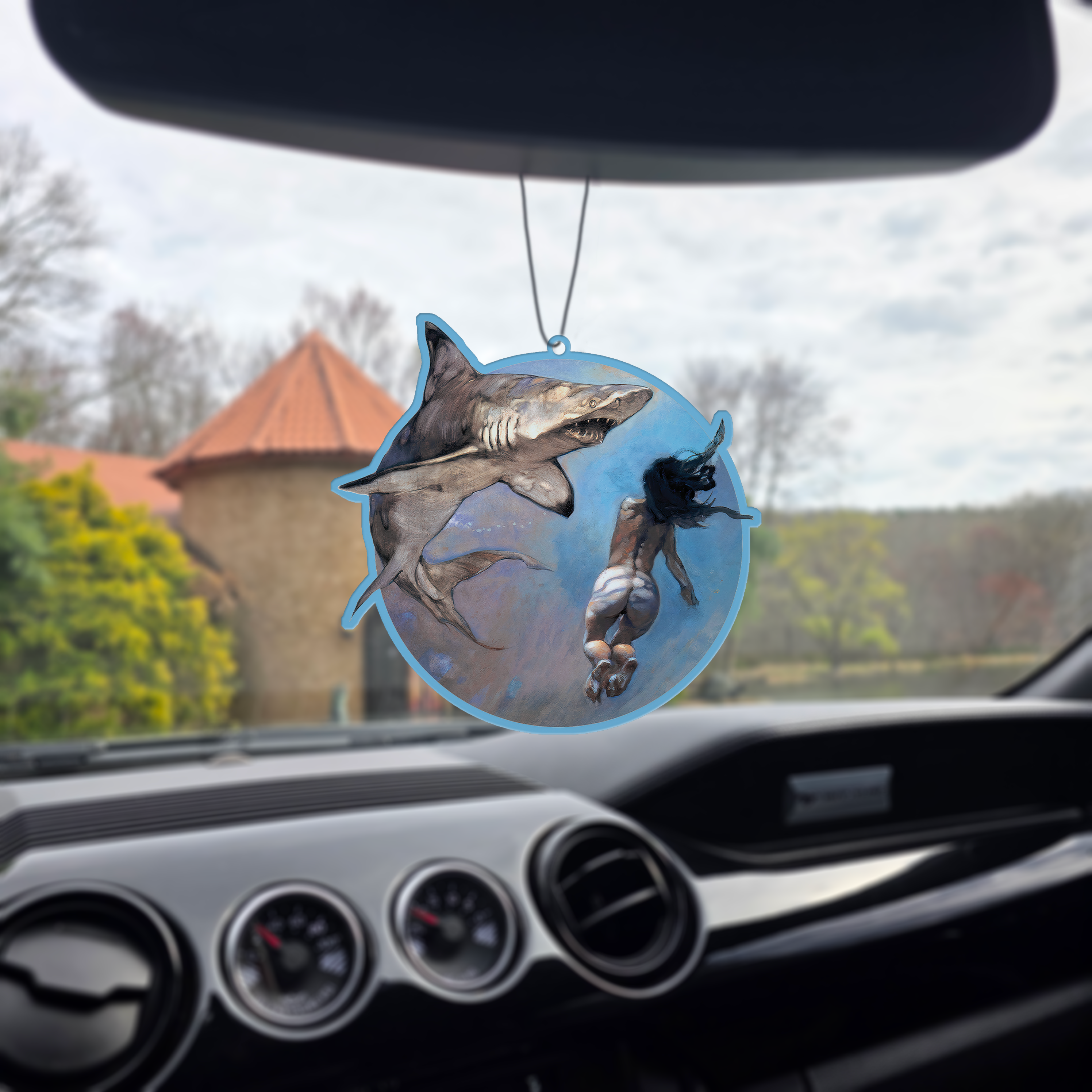 Photo of the Frazetta Requiem of a Shark air freshener hanging in a car with black interior. The Frazetta Art Museum sits in the background with gloomy clouds overhead.
