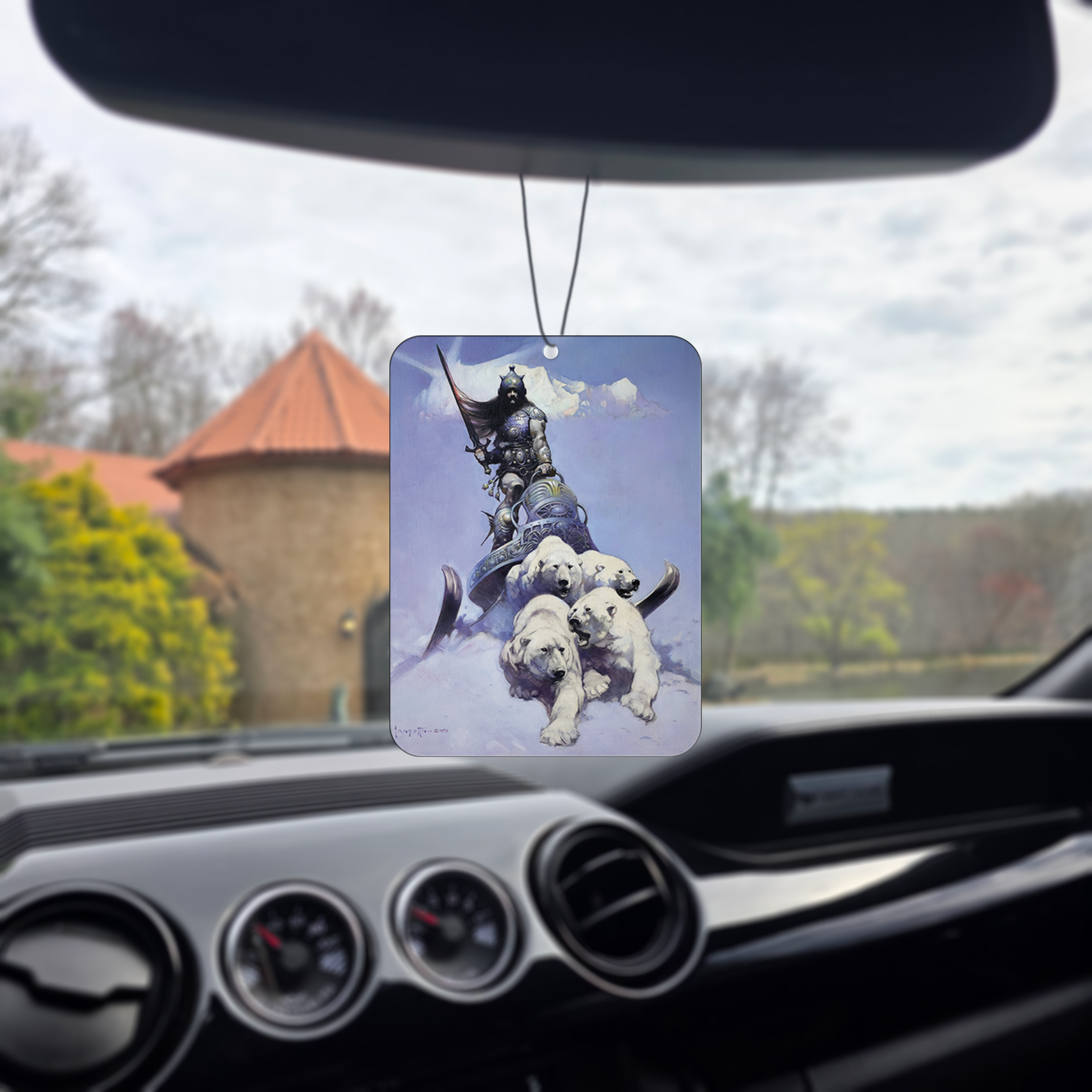 Photo of the Frazetta Silver Warrior air freshener hanging in a car with black interior. The Frazetta Art Museum sits in the background with gloomy clouds overhead.