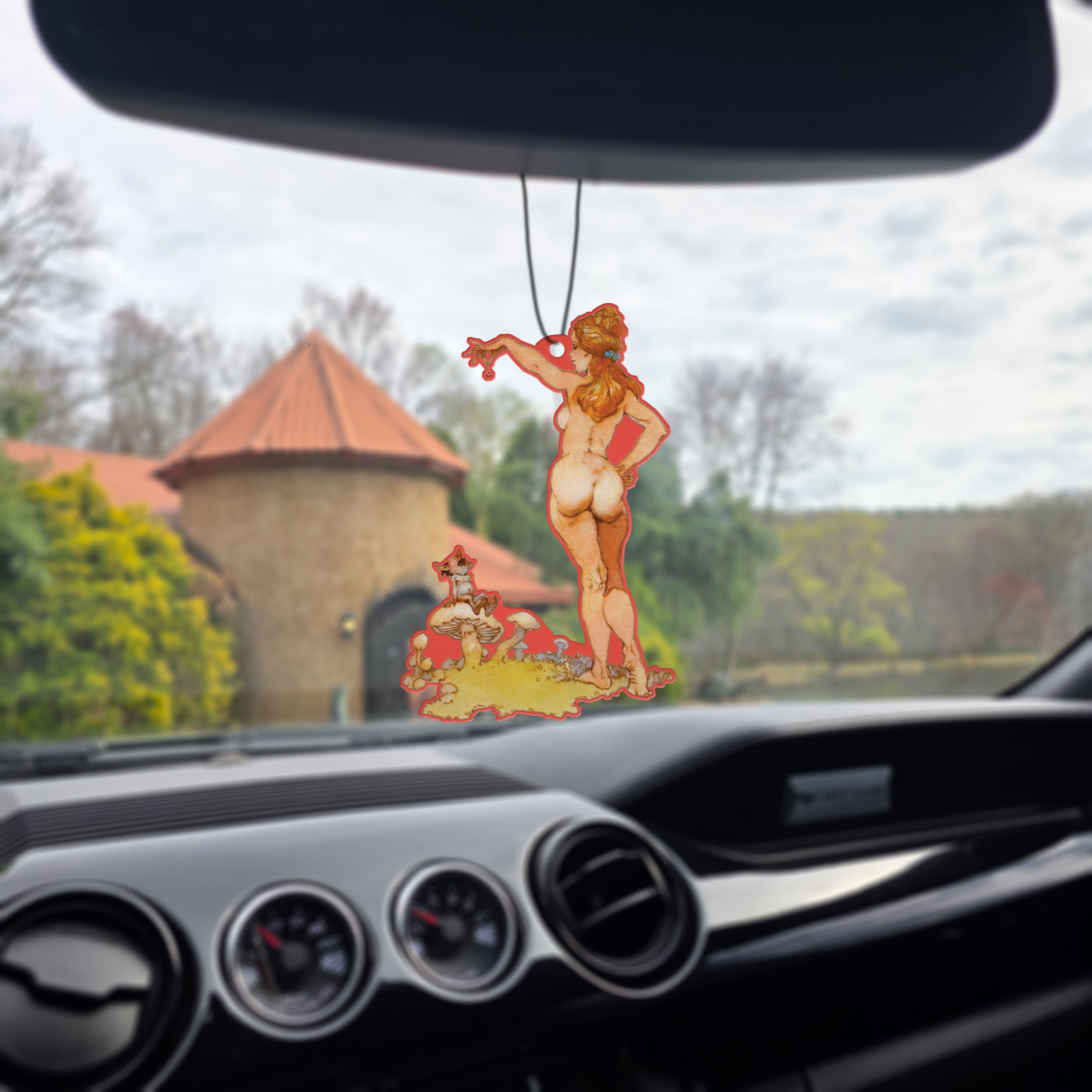 Photo of the Frazetta Teaser air freshener hanging in a car with black interior. The Frazetta Art Museum sits in the background with gloomy clouds overhead.