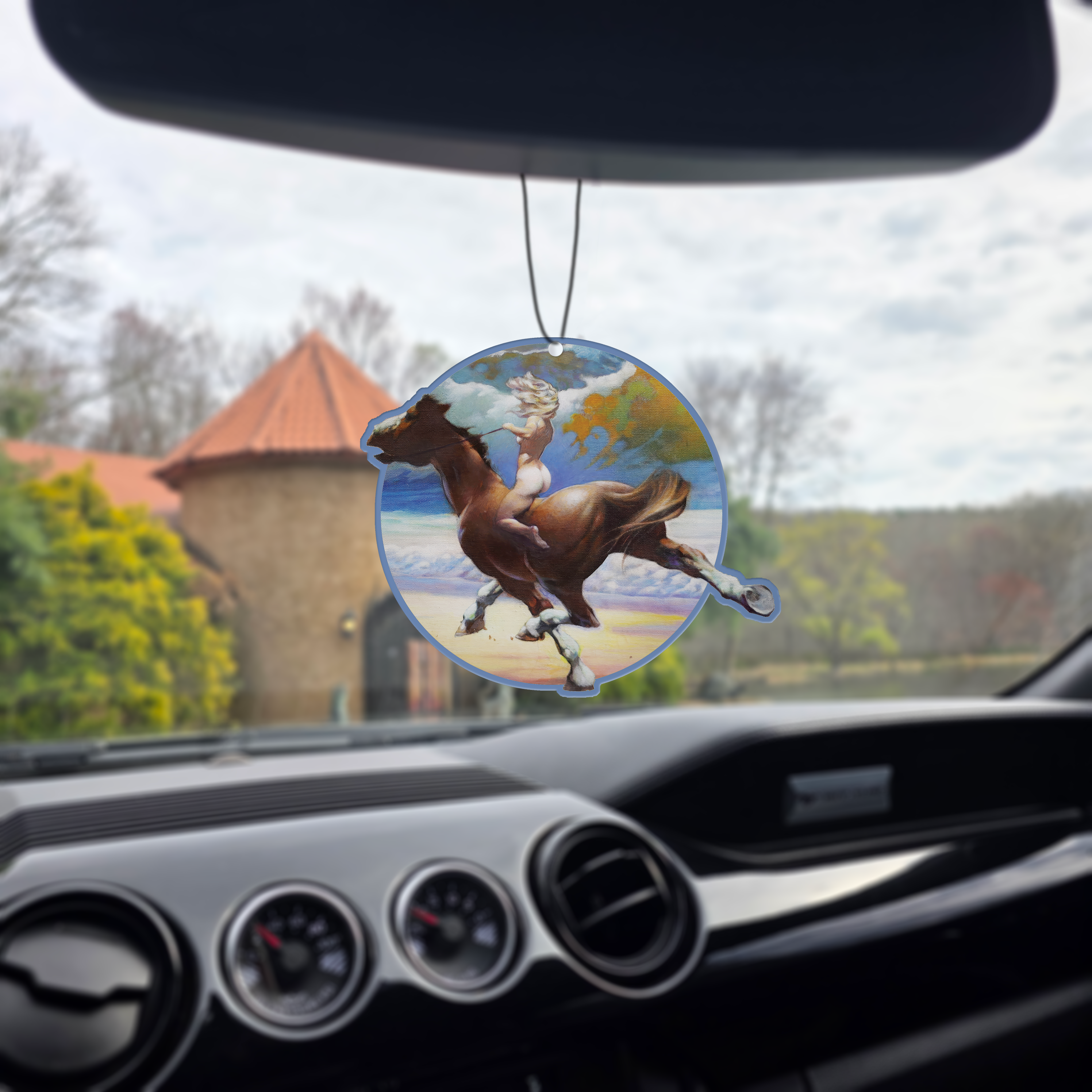 Photo of the Frazetta Wild Ride air freshener hanging in a car with black interior. The Frazetta Art Museum sits in the background with gloomy clouds overhead.