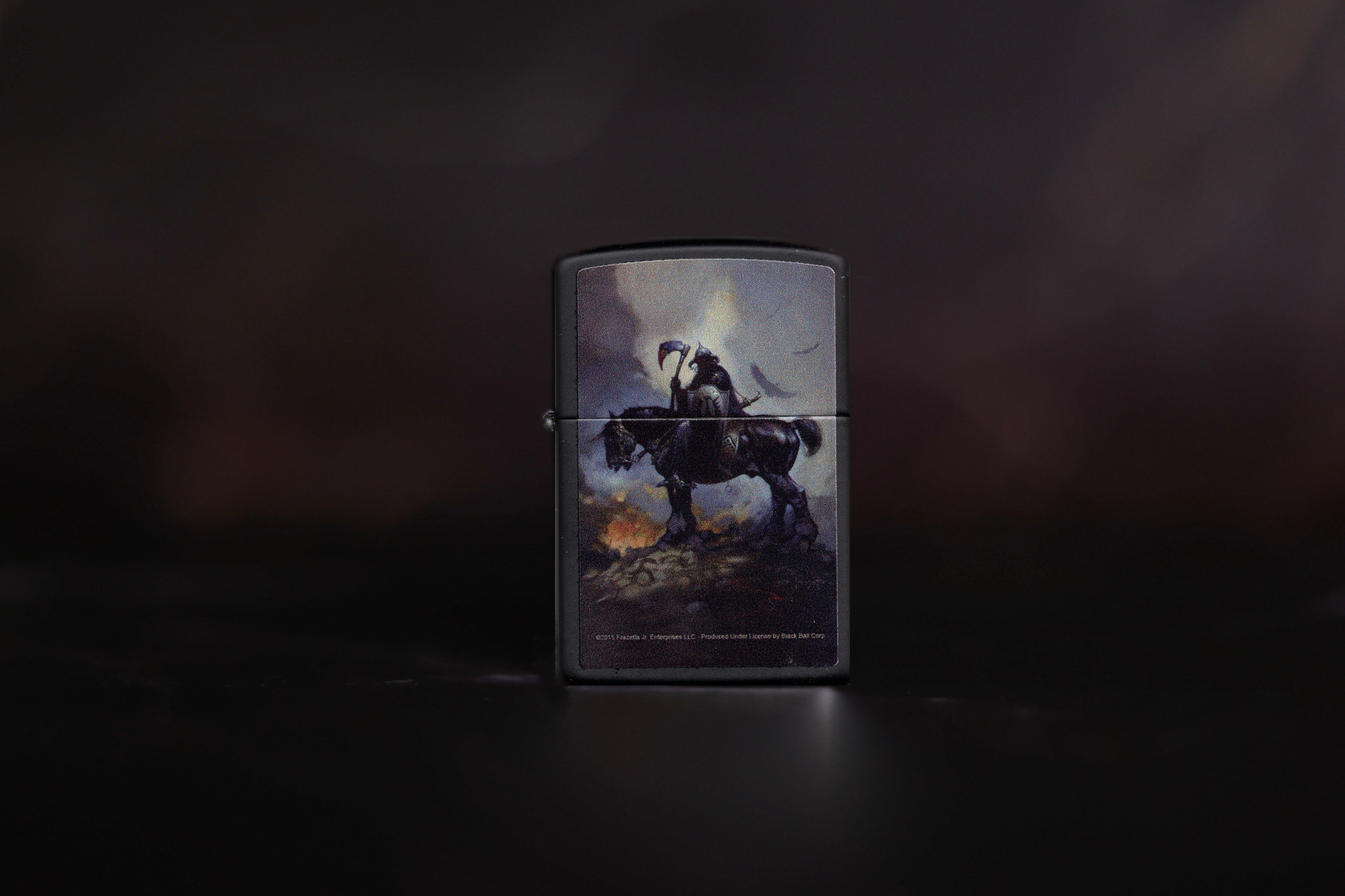 A matte black Zippo lighter featuring Frank Frazetta’s iconic “Death Dealer” painting. The artwork depicts a dark, armored warrior on horseback holding a bloodied axe, surrounded by smoke and fire. The background is atmospheric and moody, with a dramatic light piercing the stormy sky. Small copyright text appears at the bottom of the lighter, reading “©2015 Frazetta Jr. Enterprises LLC. Produced under license by Beads Bell Corp.” The lighter is centered against a dark, blurred background.