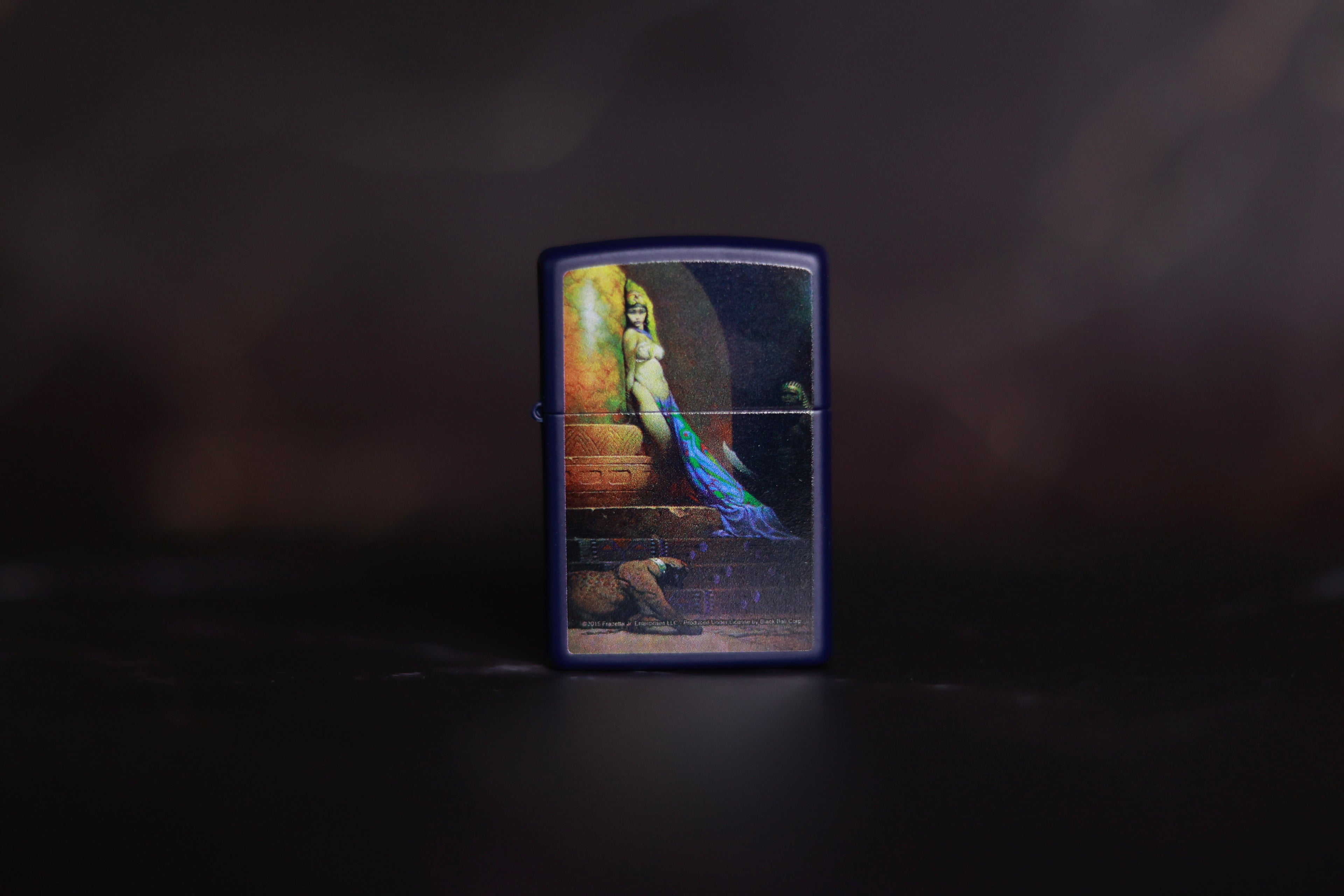 Dark blue Zippo-style lighter featuring Frank Frazetta’s Egyptian Queen artwork in full color, showing a partially draped woman standing near a stone wall with a panther at her feet, against a dark, moody background.