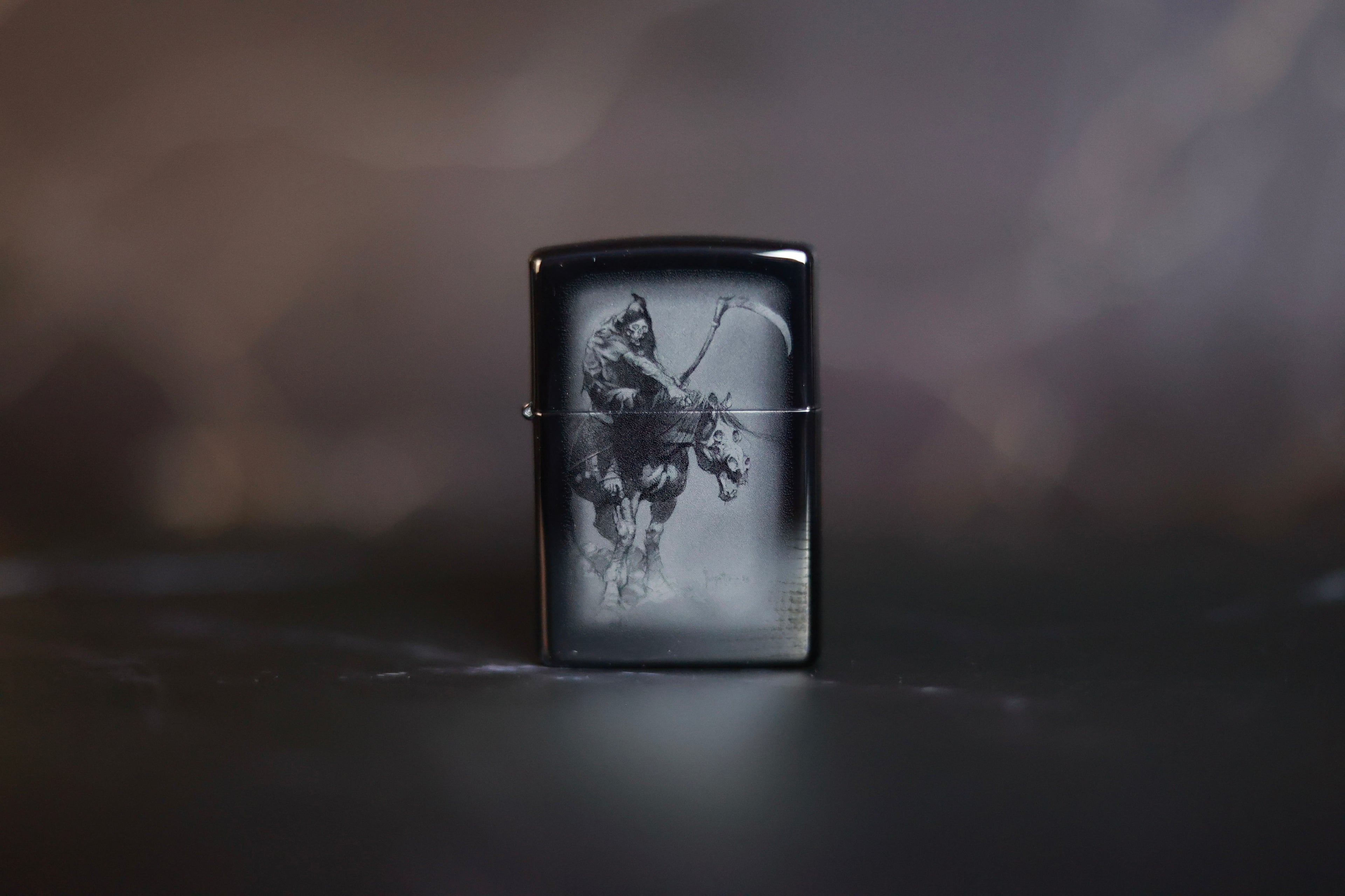 Black Zippo-style lighter featuring Frank Frazetta’s Grim Reaper artwork etched in grayscale, displayed upright on a dark, blurred background.