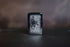 Black Zippo-style lighter featuring Frank Frazetta’s Grim Reaper artwork etched in grayscale, displayed upright on a dark, blurred background.
