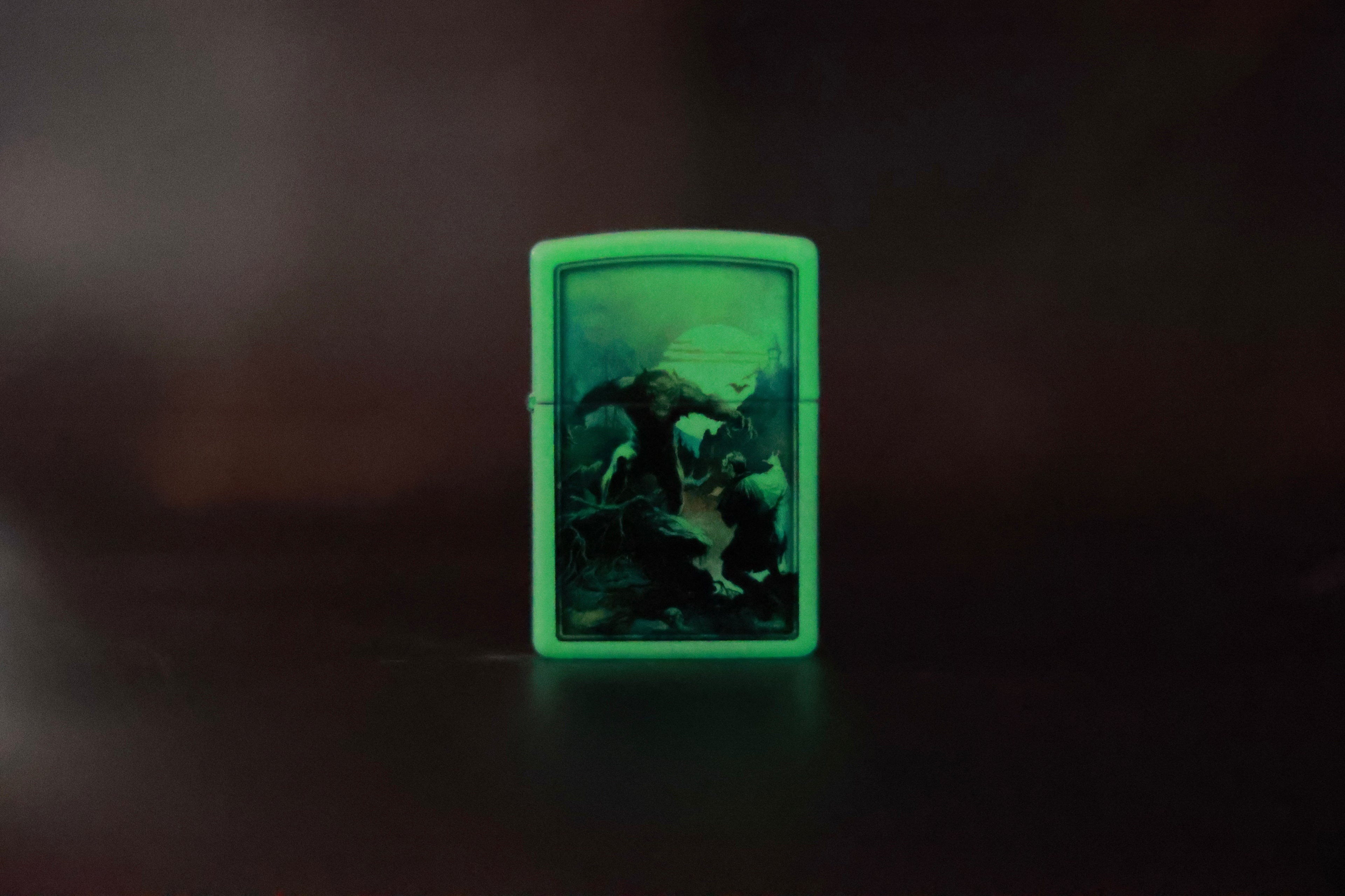 Same Wolfmoon Zippo-style lighter shown glowing in the dark, with the eerie green illumination highlighting the werewolf scene under the moonlight.