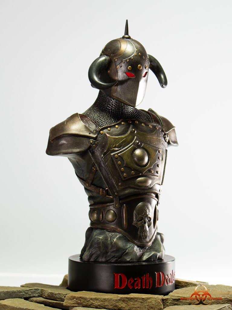 A front-facing view of the 1/4 scale Death Dealer bust, featuring a horned helmet, glowing red eyes, and battle-worn armor with intricate detailing. The statue is mounted on a black base with "Death Dealer" inscribed in bold red lettering, set on a stone-like surface.
