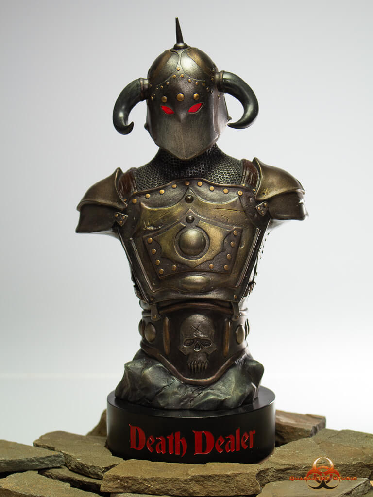 A front-facing view of the 1/4 scale Death Dealer bust, showcasing its horned helmet, glowing red eyes, and battle-worn armor. The intricate details of the chest plate and skull emblem on the base are prominently displayed, with the "Death Dealer" inscription in bold red lettering on the stand. The bust rests on a stone-like platform.