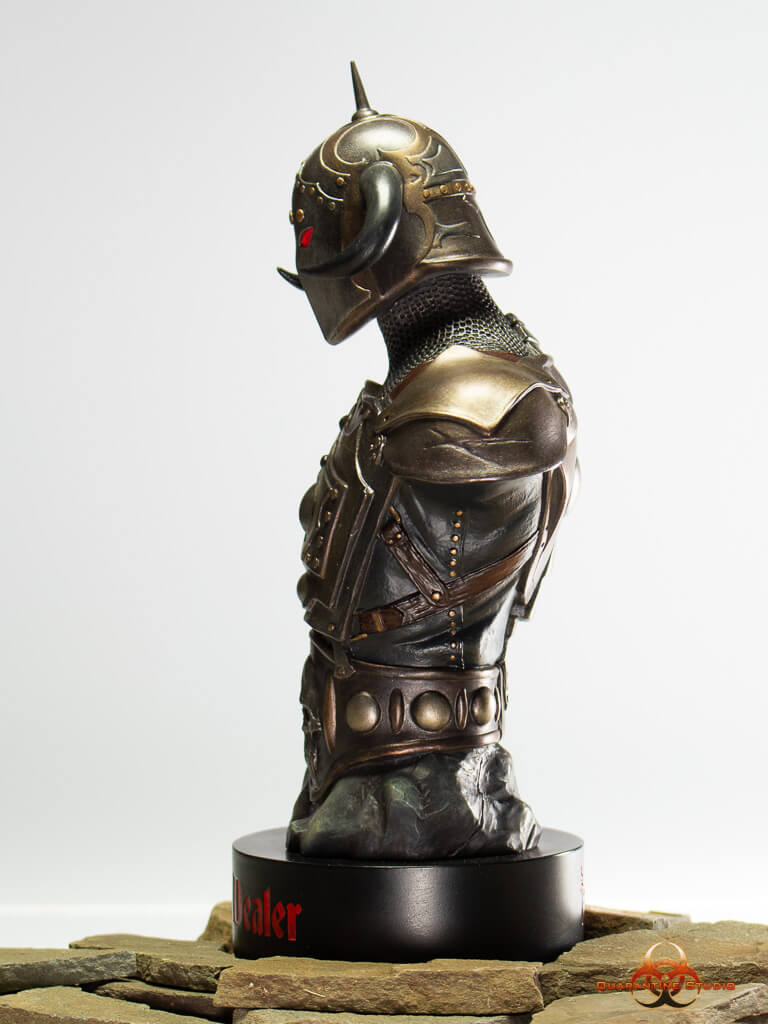 A side profile view of the 1/4 scale Death Dealer bust, highlighting the intricate details of the horned helmet, glowing red eyes, and layered armor. The statue is mounted on a black base with red lettering, resting on a stone-like surface.