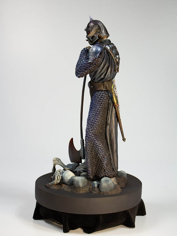 Side view of the Death Dealer 1:6 scale statue by Quarantine Studio, highlighting intricate chainmail armor, a sheathed sword with gold accents, and a battle-worn axe resting among skeletal remains. The warrior&