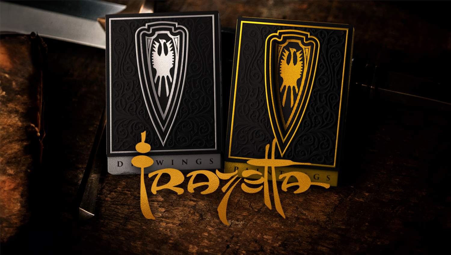 Two elegantly designed playing card decks rest on a rustic wooden surface. One deck features a black and silver design, while the other is black and gold, both adorned with an intricate shield emblem depicting a stylized eagle. 